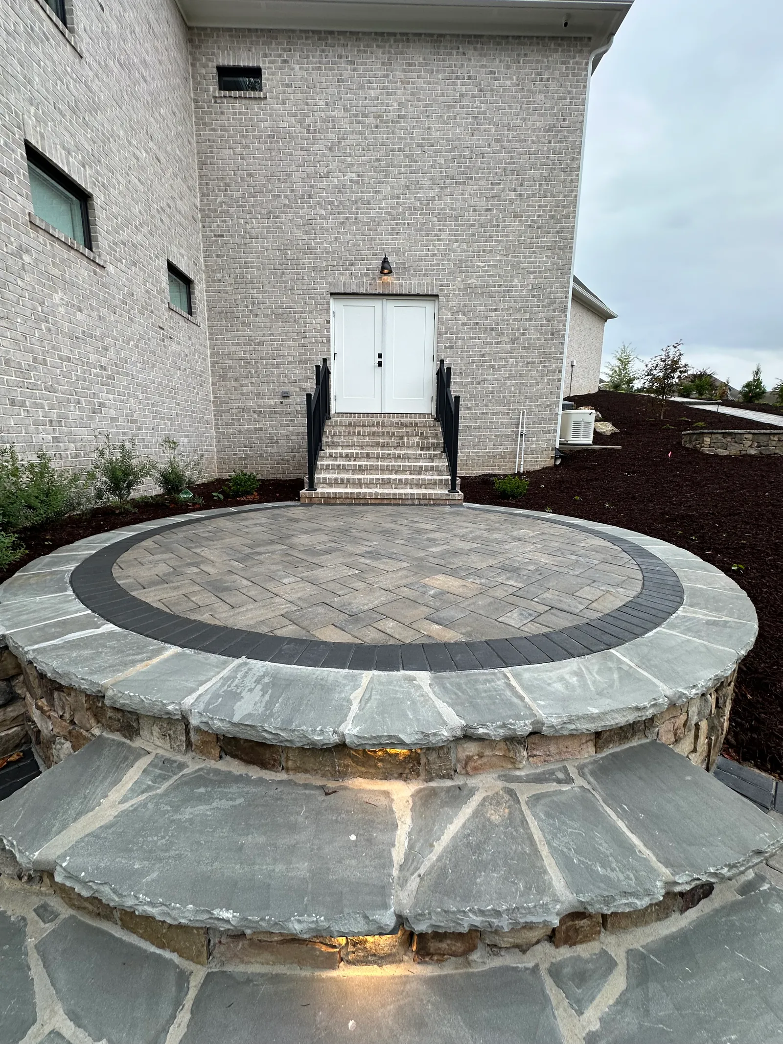 Circular herringbone paver patio with stone steps and a retaining wall leading to a home entrance