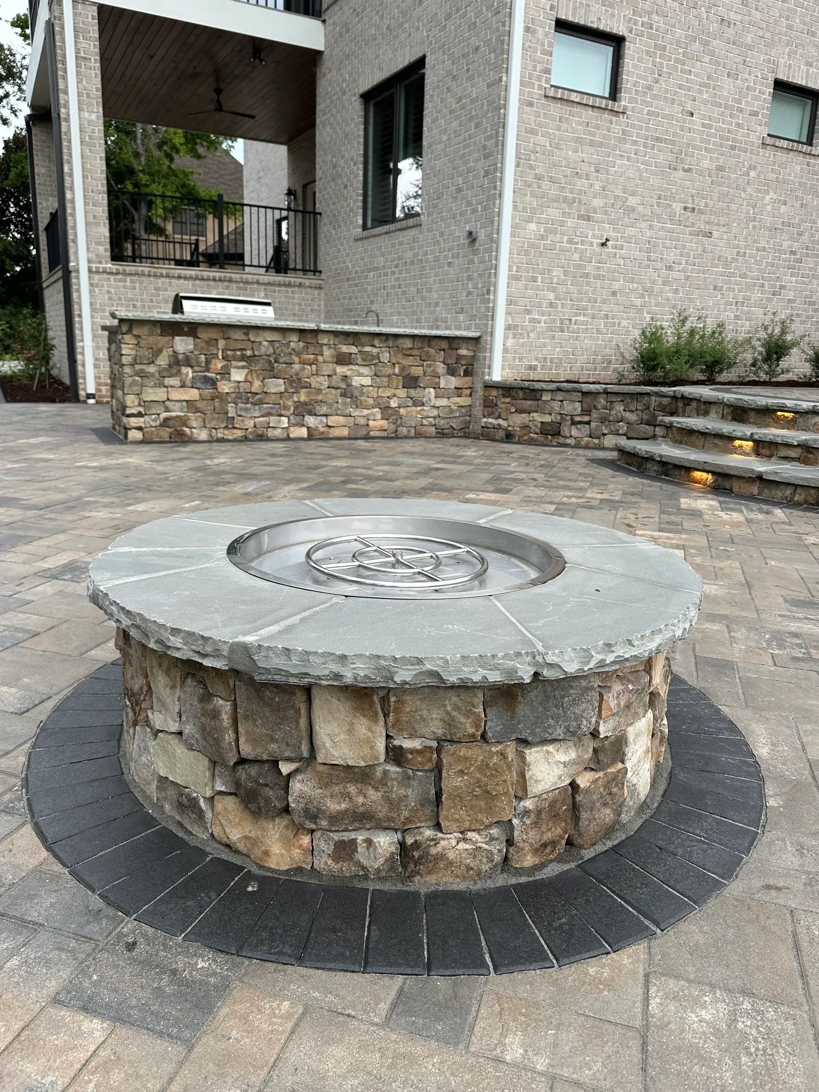 Round natural-stone fire pit with a gas insert on a paver patio, outdoor kitchen visible in the background