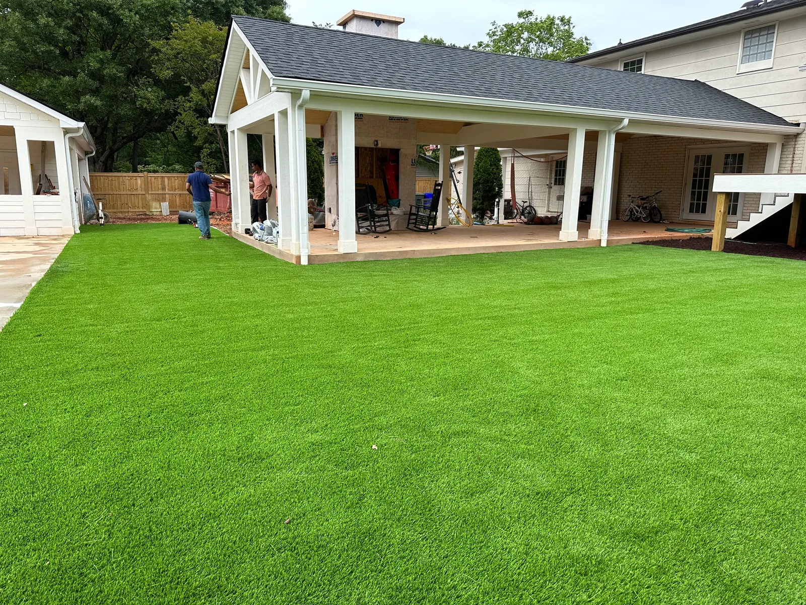 Wide artificial turf lawn installation beside a covered outdoor pavilion, installation crew finishing the seams